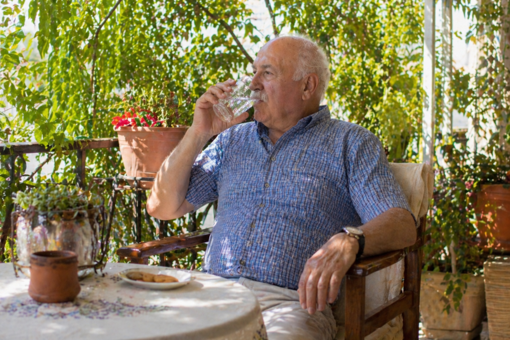 Έλληνας ηλικιωμένος κάθεται σε σκιερό μπαλκόνι το καλοκαίρι και πίνει νερό