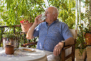 Έλληνας ηλικιωμένος κάθεται σε σκιερό μπαλκόνι το καλοκαίρι και πίνει νερό